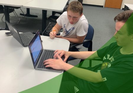 Two students working on creating content with laptops in the library with a green swoosh in the lower right corner