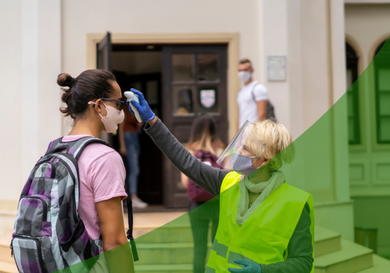 Student with a mask on getting a temperature check from a school official in a mask and shield in front of the school building