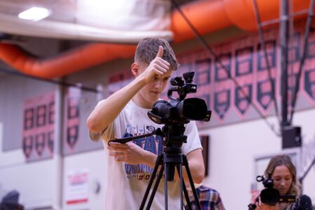 A blonde teenage boy wearing a white shirt is taking a video on a professional video camera in a school gym. He has one hand holding the camera and the other giving a thumbs up to his video subject.