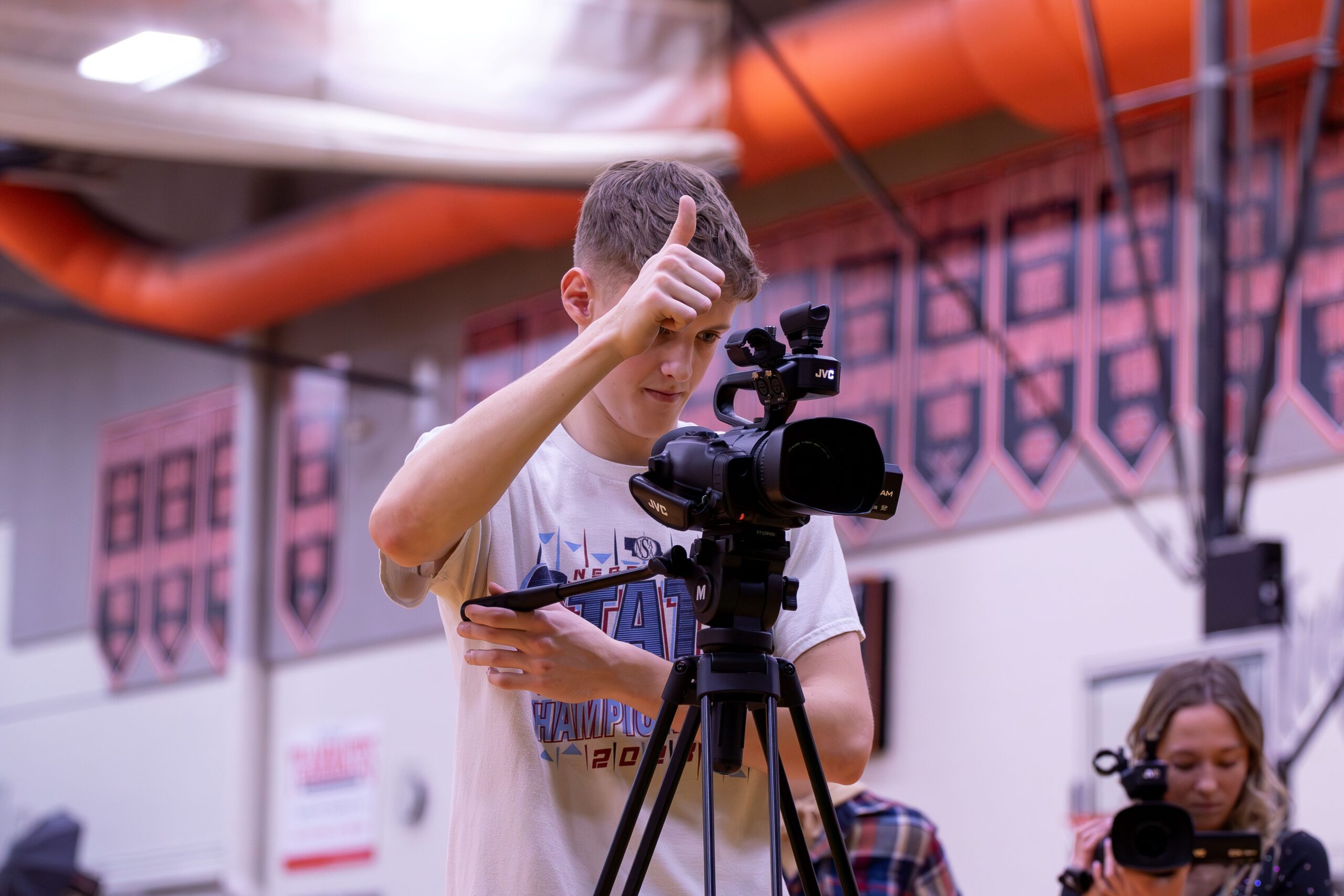 A blonde teenage boy wearing a white shirt is taking a video on a professional video camera in a school gym. He has one hand holding the camera and the other giving a thumbs up to his video subject.
