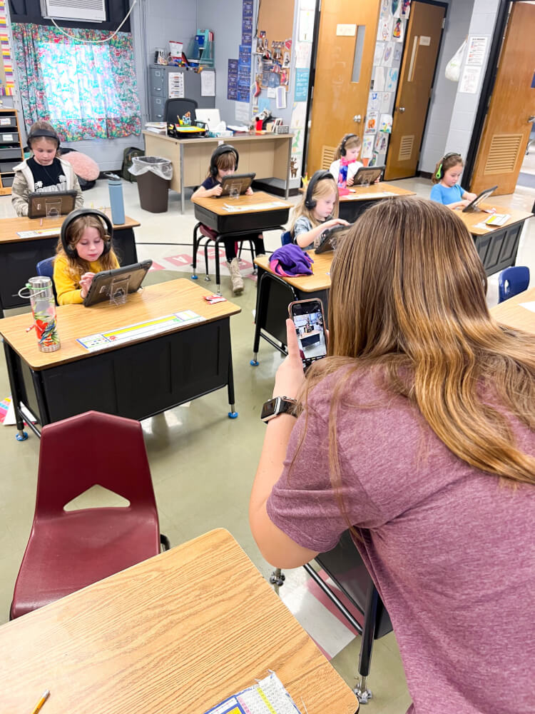 A student films video on a phone while classmates work on tablets nearby during a classroom activity.