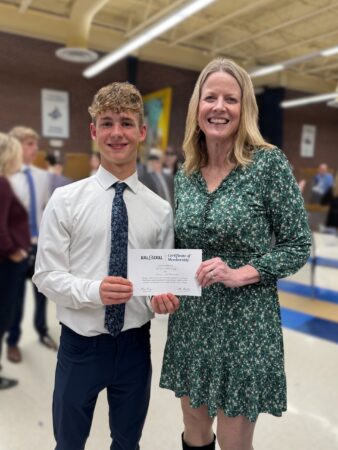 Dr. Jill Johnson poses with Drew Collings in a high school gym following an awards ceremony. They each old a side of Drew's Quill & Scroll certificate and smile at the camera. 