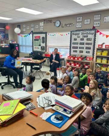 A high school basketball player sits at the front of an elementary classroom holding a book. She and all the elementary students smile at the camera to their left. 