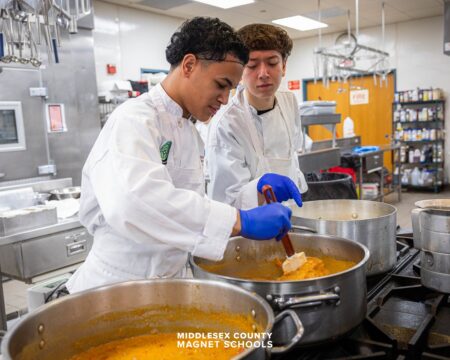 Two students work together on a culinary creation in a large pot. They are in an industrial looking kitchen and garbed in professional culinary attire. 