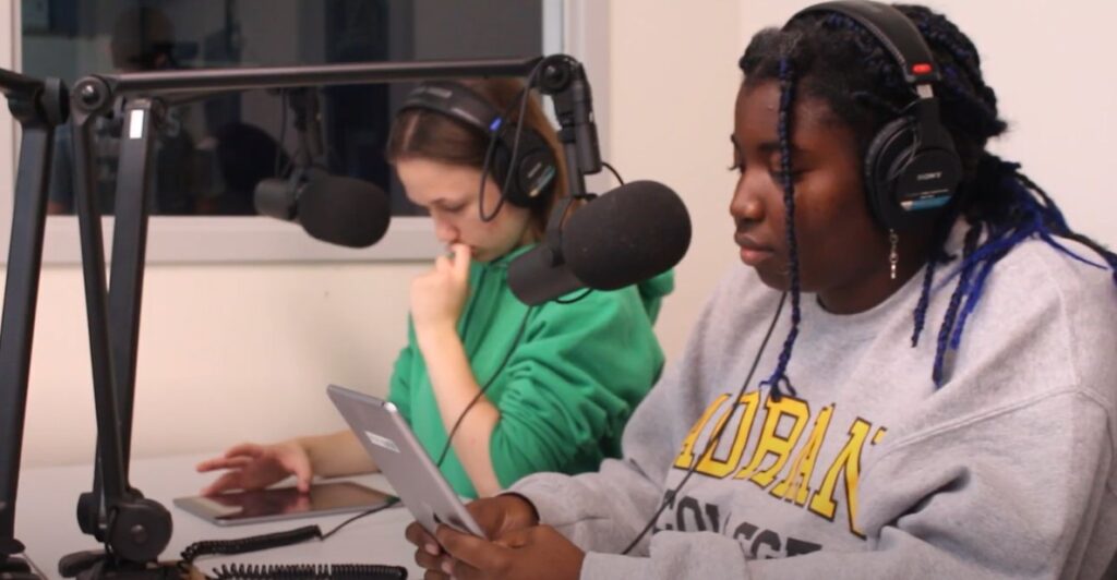 Two students sit at table with podcast-style microphones, wearing over the ear headphones. They are both reviewing notes on an iPad. 