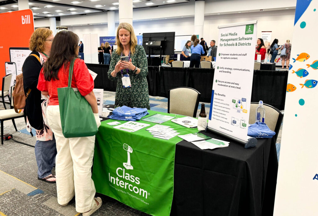 Dr. Jill Johnson stands behind the Class Intercom table at FACTS Elevate. She is showing two women, in front of the table, how to use Class Intercom on her phone. 