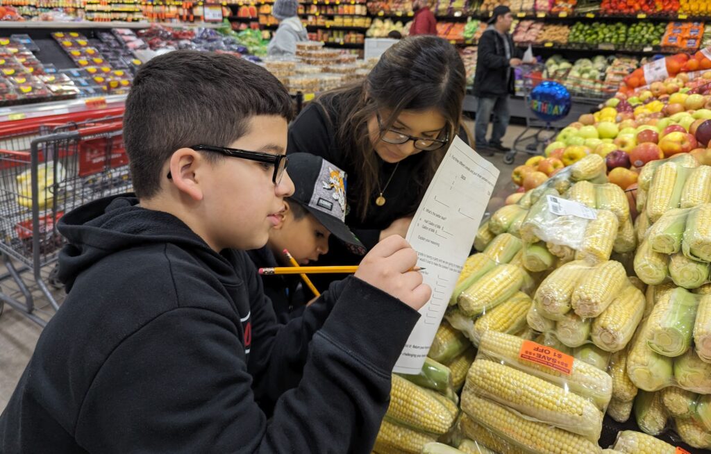 A woman and two young boys stand in the vegetable section of a grocery store. The boys are using pencils to write on a sheet of paper. 