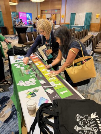 Two women at a conference table with a green tablecloth, writing on cards surrounded by colorful items like stickers and markers. One woman has a beige tote bag and the other has short blonde hair and glasses. The room has chairs, a coffee cup, and booths with attendees in the background.