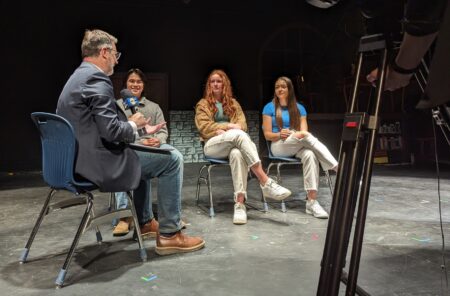 Three high school students being interviewed by the local news on their auditorium stage. 