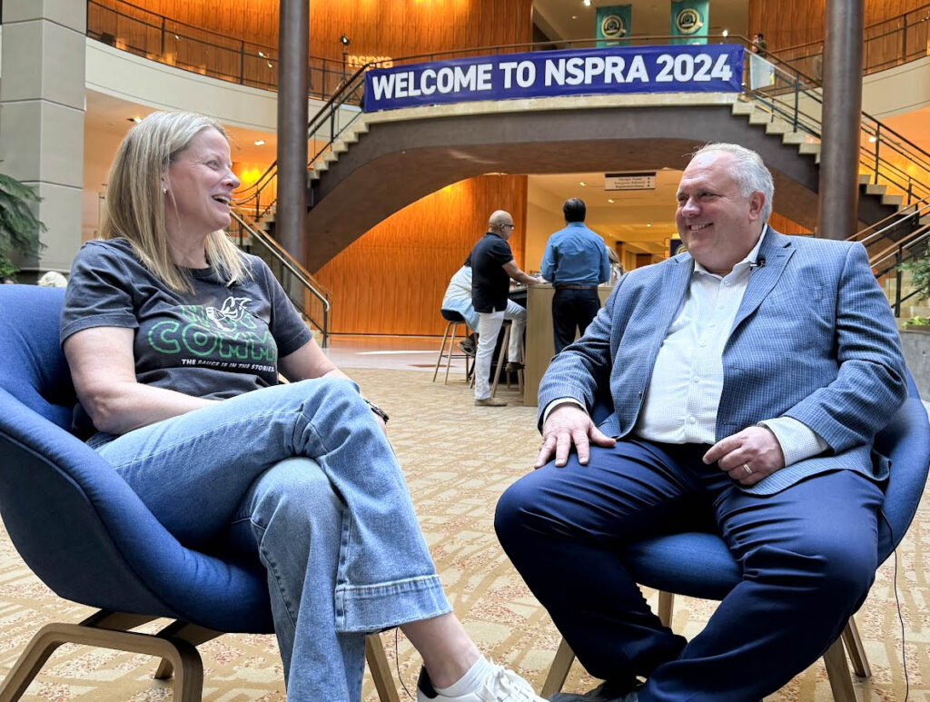 Dr. Jill Johnson and Teddy Gabbart talk and laugh in two blue chairs under a blue sign that reads, "Welcome to NSPRA 2024". 