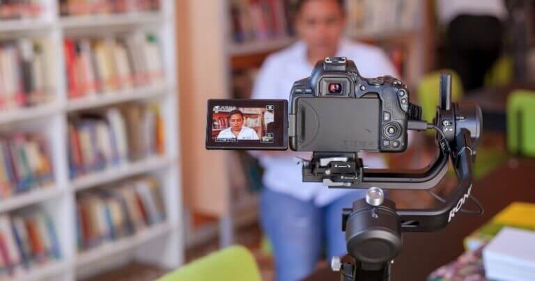 Close-up of a DSLR camera recording a student in a school library, with the subject in focus on the camera screen and bookshelves blurred in the background.