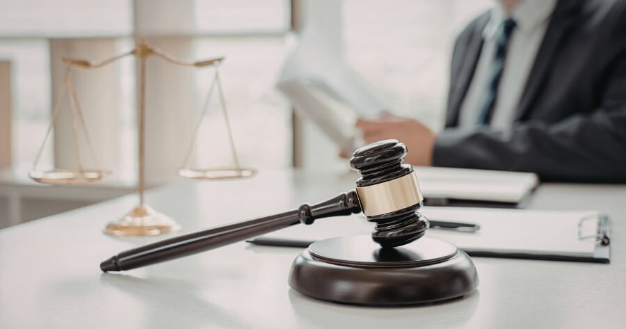 A gavel and scale sit on a desk with clipboards and papers. There's a person in a suit sitting at the desk reading a document.