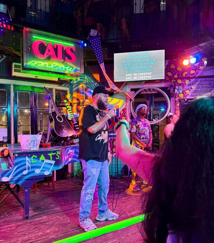 A person singing into a microphone on a neon-lit karaoke stage with lyrics on a screen behind them.