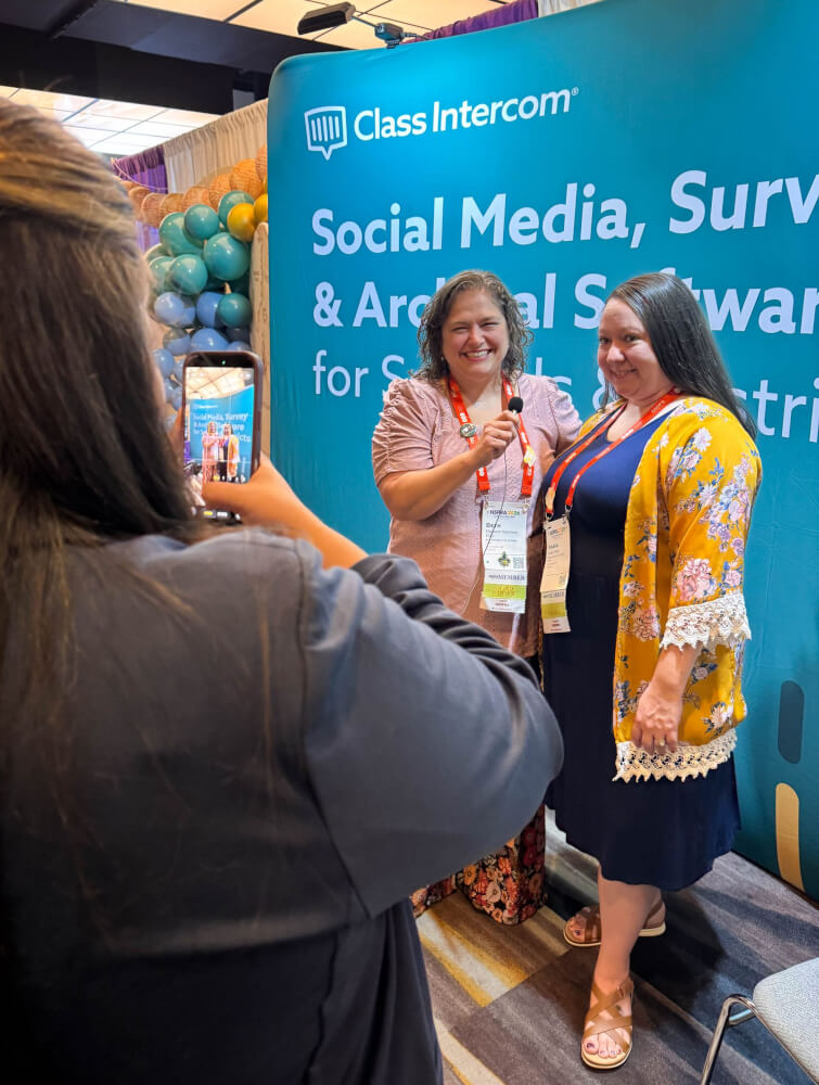 Two conference attendees with award ribbons being recorded on a phone in front of a Class Intercom booth backdrop.