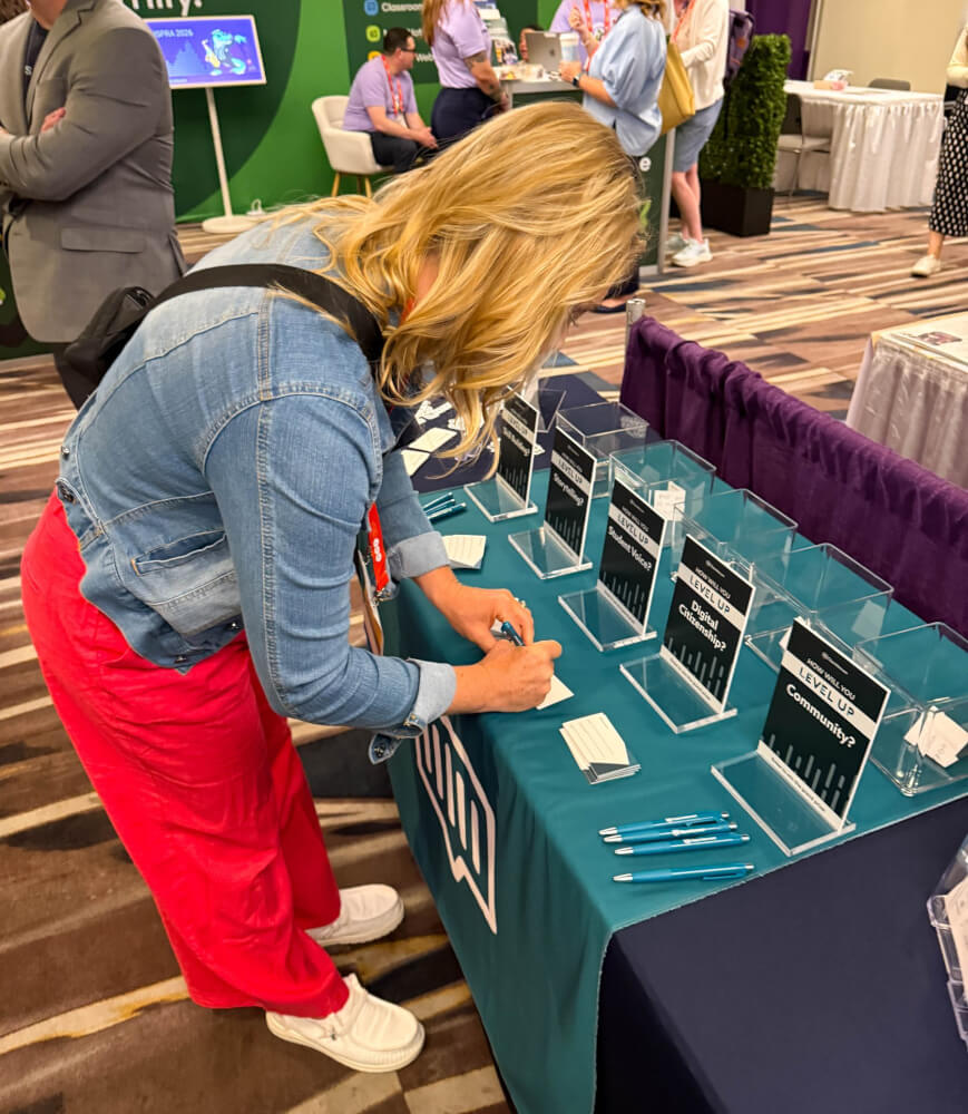 A conference attendee writing on a card at the Class Intercom booth, beside signs reading How will you level up: Storytelling, Student Voice, Digital Citizenship, Community.