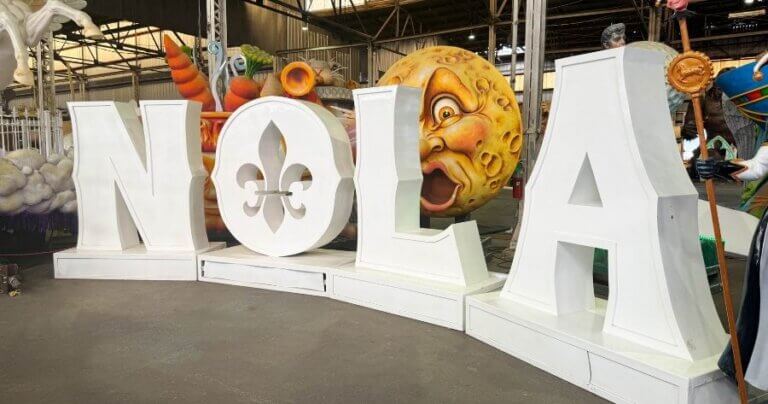 Large white NOLA letters with a fleur-de-lis on display among Mardi Gras floats in a New Orleans warehouse.