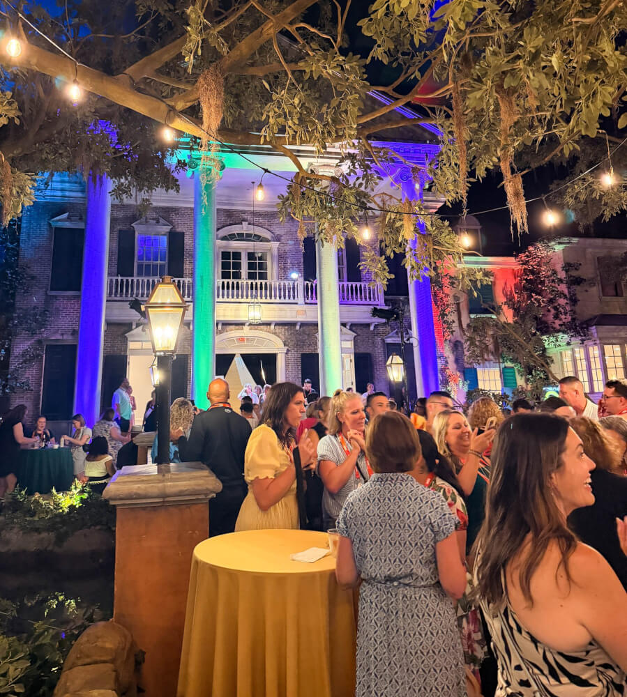 Conference attendees talking outdoors at night beneath string lights, with a columned building lit in blue and green behind them.